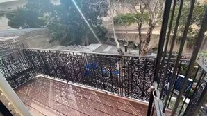 Ein Balkon mit verzierten Metallgeländern, einem Holzboden und Blick auf einen Baum sowie ein Gebäude im Hintergrund.