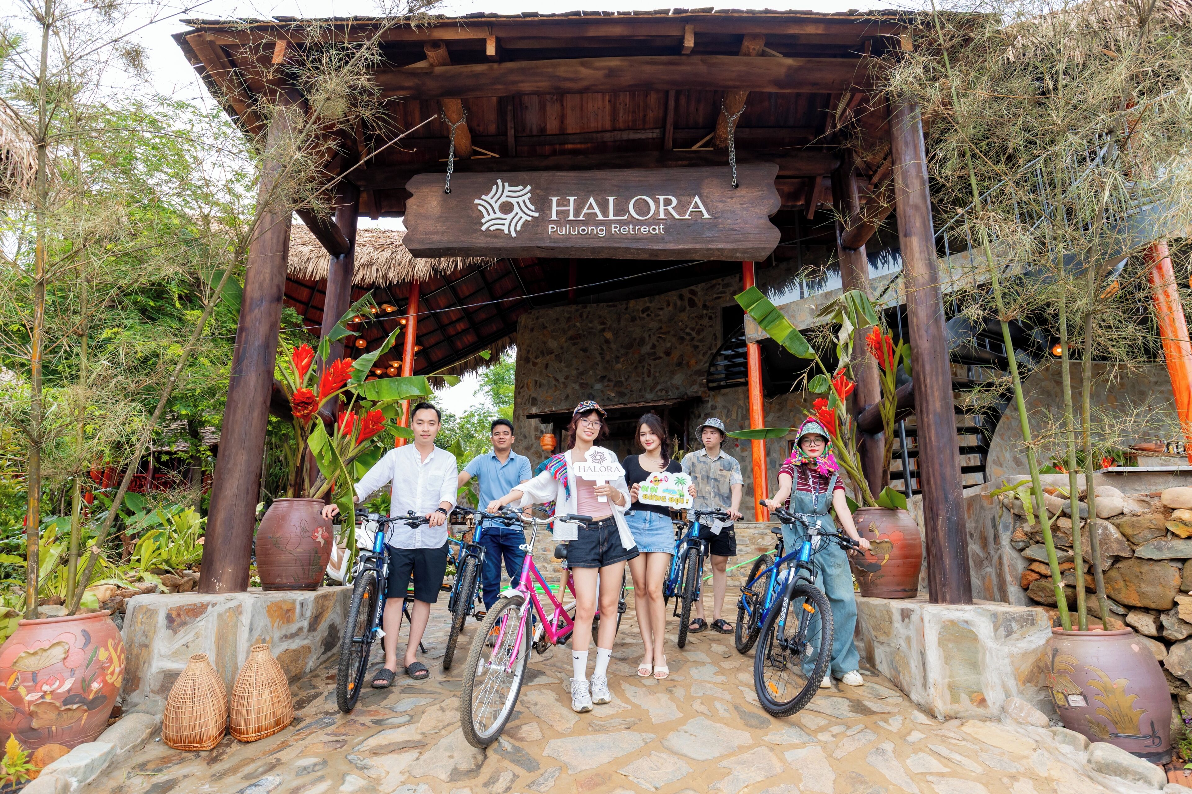 Una estructura de madera con un letrero que dice 'HALORA Puluwong Retreat', rodeada de plantas en macetas y bicicletas.