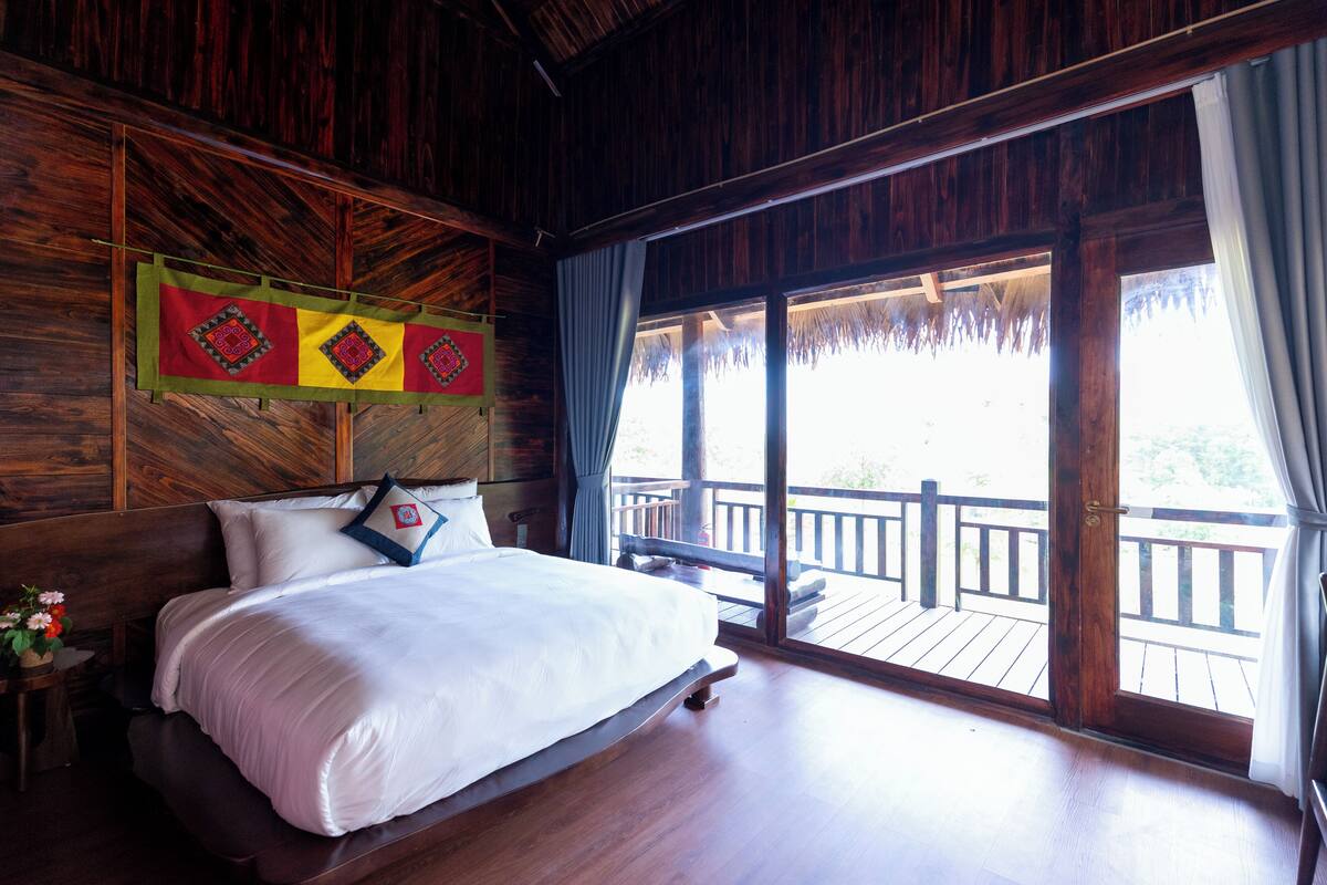 A wooden room with a bed, a window with curtains, and a view of a balcony.