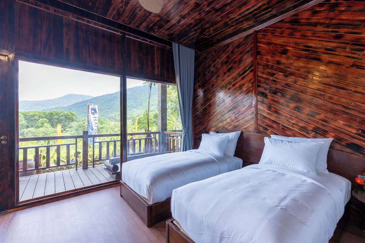 A room with two beds, a balcony with a wooden railing, and a view of greenery and mountains.