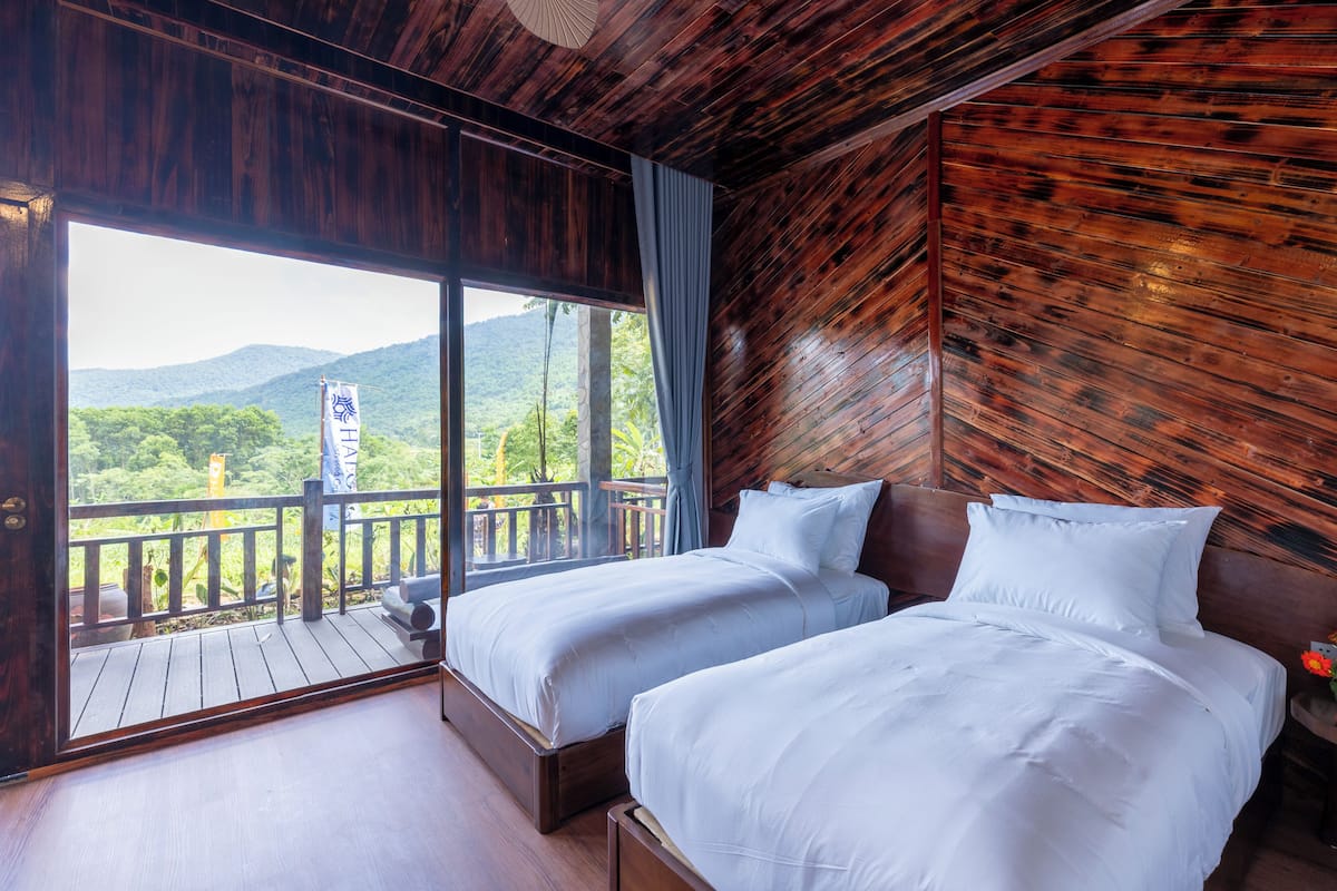 Ein Zimmer mit zwei Betten, einem Balkon mit Holzgeländer und Blick auf Grünflächen und Berge.