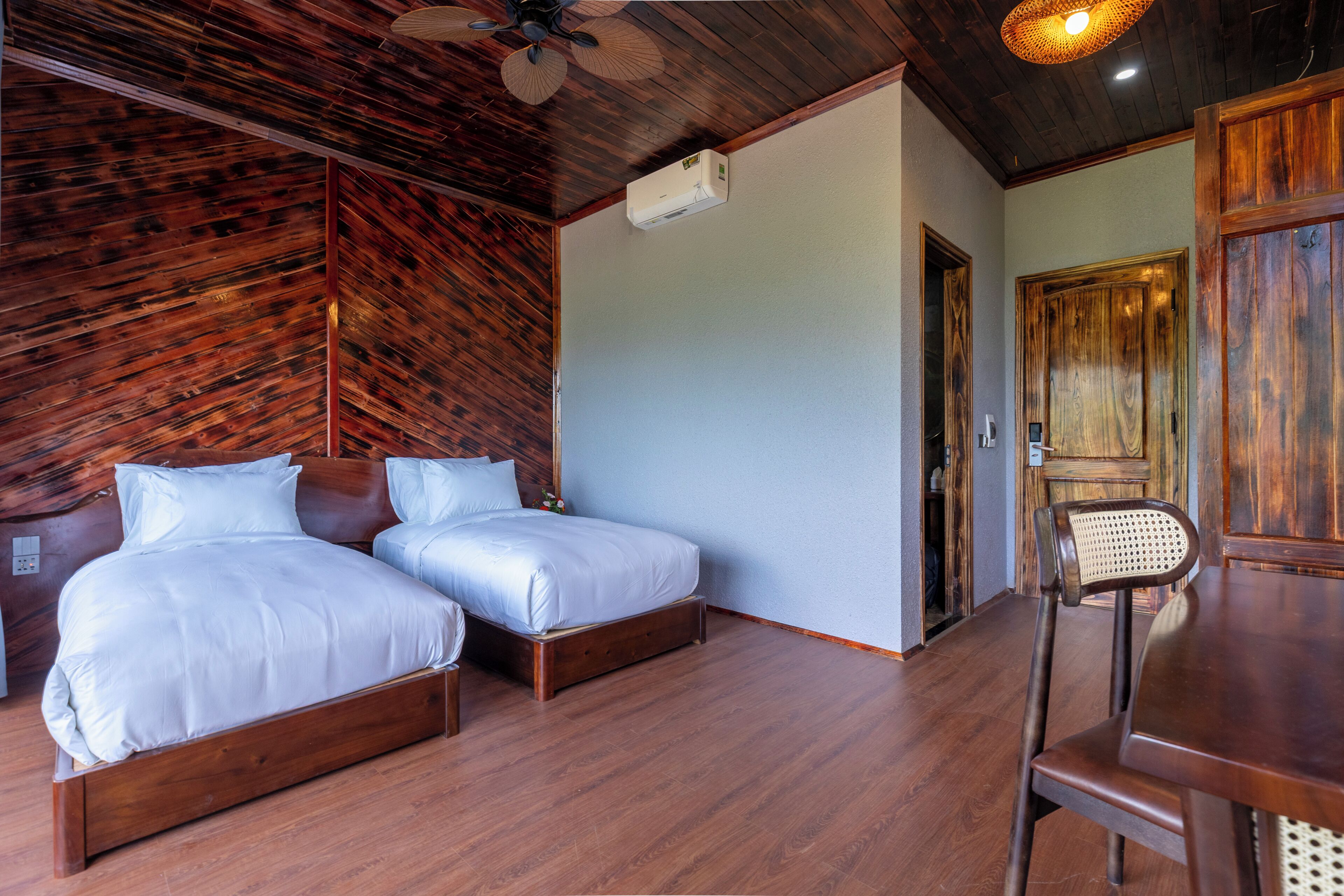 A room with two wooden beds, a ceiling fan, a wooden table, and a door.