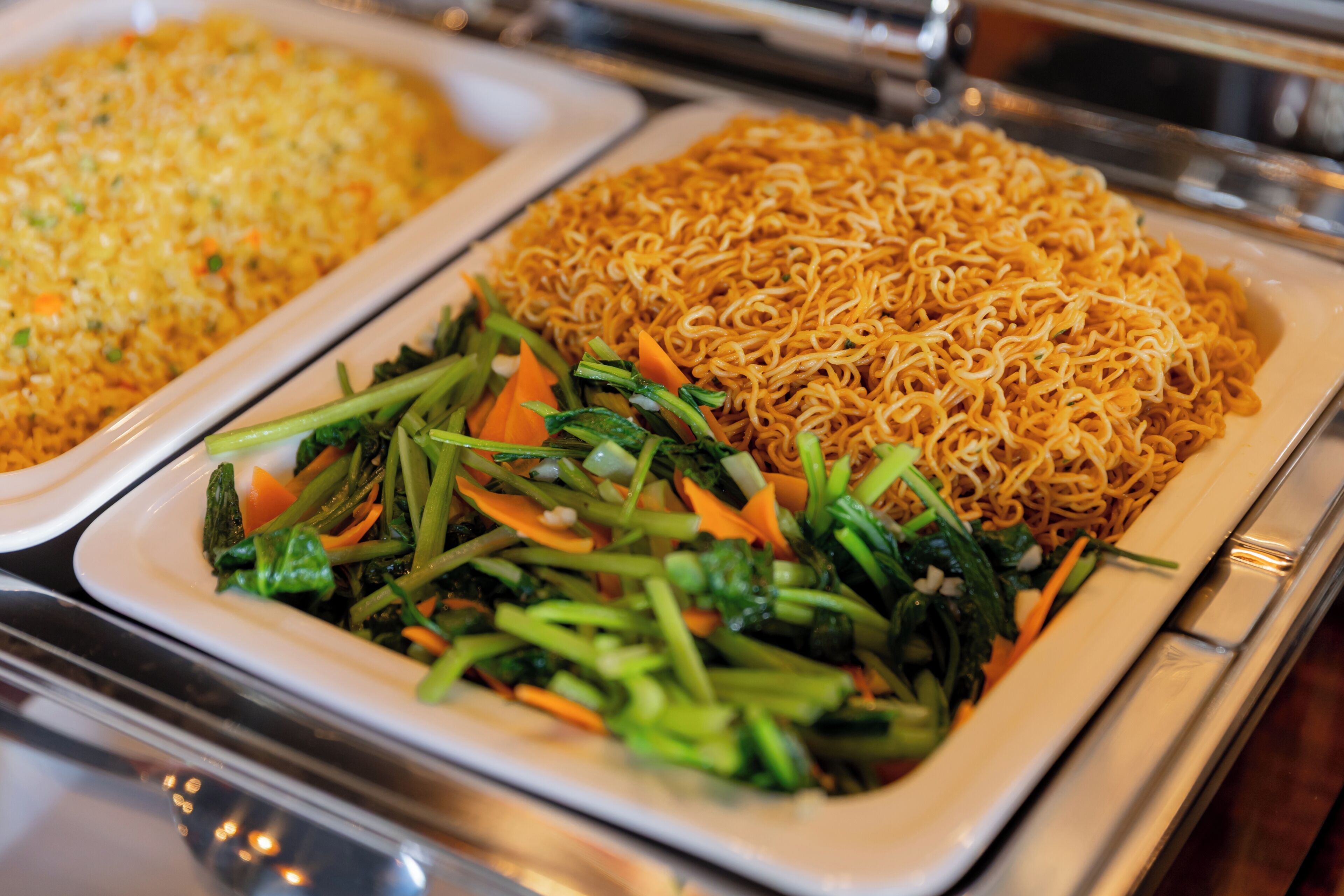 Un bufé con diversos platos, incluyendo fideos, arroz frito y un salteado de verduras.