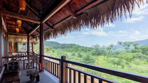 A wooden deck with a thatched roof, a fire extinguisher, and a view of a green landscape. - Halora Puluong Retreat (Pu Luong)