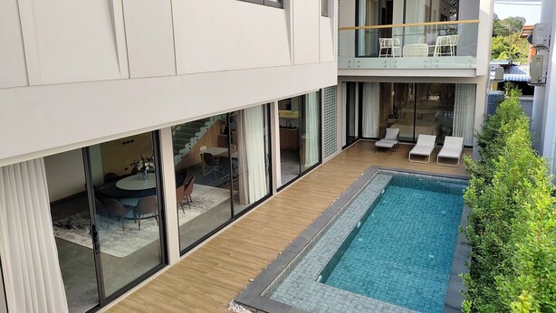 Ett modernt hus i två våningar med en swimmingpool, glasdörrar och en balkong med solstolar.
