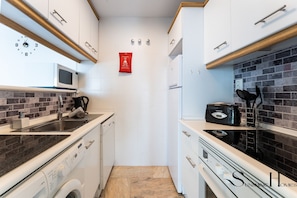 Eine kompakte Küche mit weißen Schränken, einer Mikrowelle, einem Toaster und einer Kaffeemaschine. An der Wand hängt ein rotes Schild und die Wand ist mit Fliesen verkleidet.
