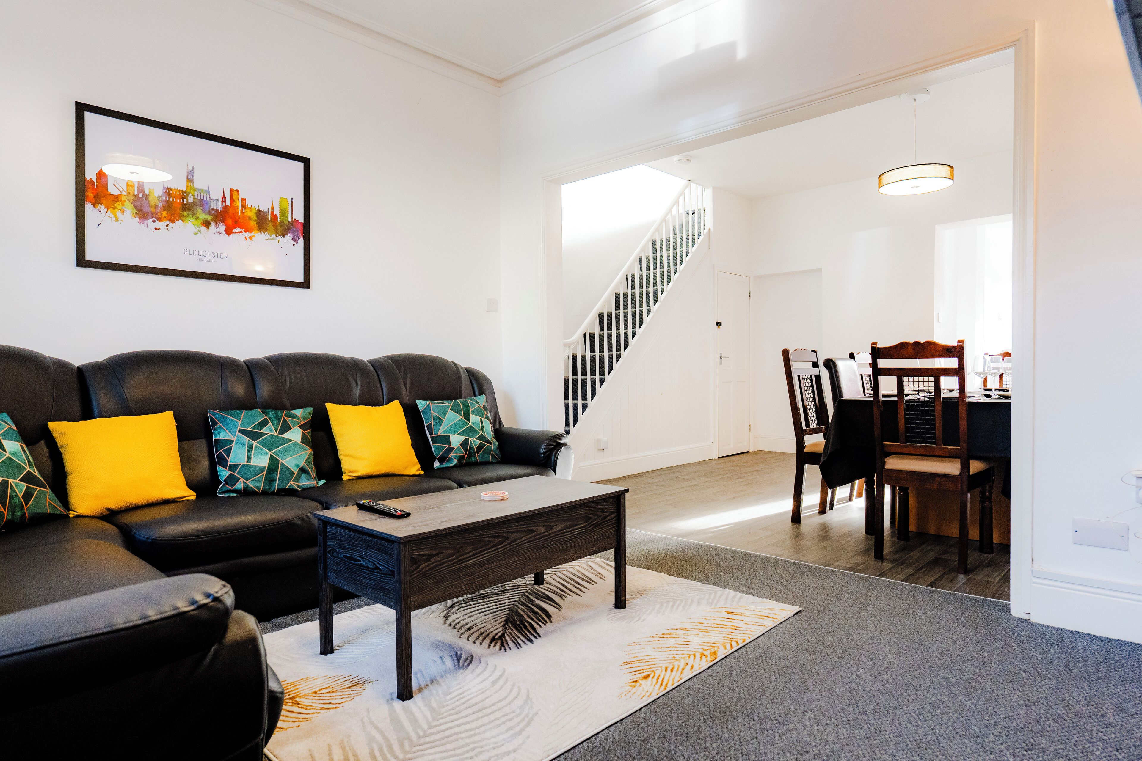 A living room with a black leather sofa, colorful cushions, a coffee table, and a cityscape artwork on the wall.