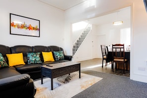 A living room with a black leather sofa, colorful cushions, a coffee table, and a cityscape artwork on the wall. - Kingsholm House - 4 Bedrooms - On the Rainbow Street (Gloucestershire)