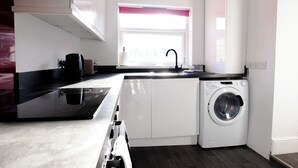 A compact kitchen with a washing machine, a coffee maker, and a window with a pink blind.