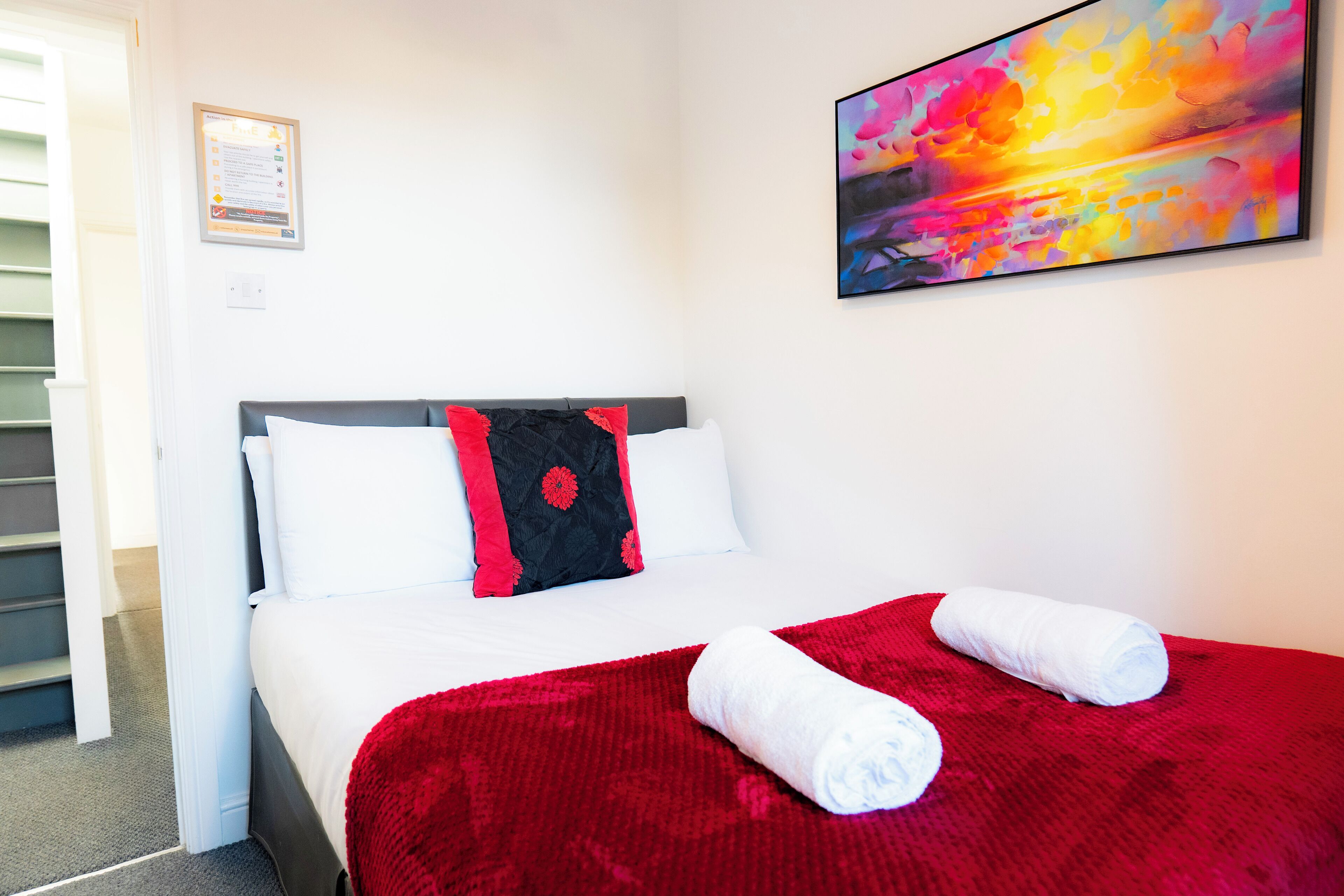 Habitació d'hotel amb llit, manta vermella, almoinades blanques i un quadre a la paret.