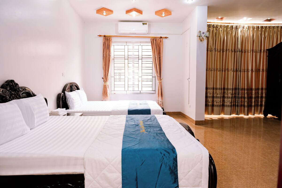 Habitación de hotel con dos camas, un ventanal grande con cortinas, un equipo de aire acondicionado y una barra para cortinas con cortinas decorativas.