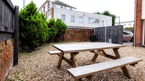A wooden picnic table in a gravel area with a brick wall and a building in the background. - Gloucester Kingsholm 6 Bedrooms (Gloucester)