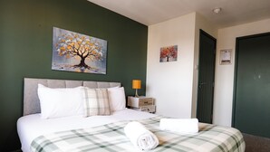 A neatly made bed with a plaid bedspread and a rolled towel, a nightstand with a lamp, and two paintings on the wall. - Gloucester Kingsholm 6 Bedrooms (Gloucester)
