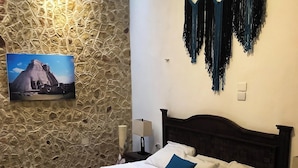 En seng med hvidt og turkis sengetøj, en træsenggavl og et natbord med en lampe. En stenvæg og et indrammet billede af en pyramide er synlige.