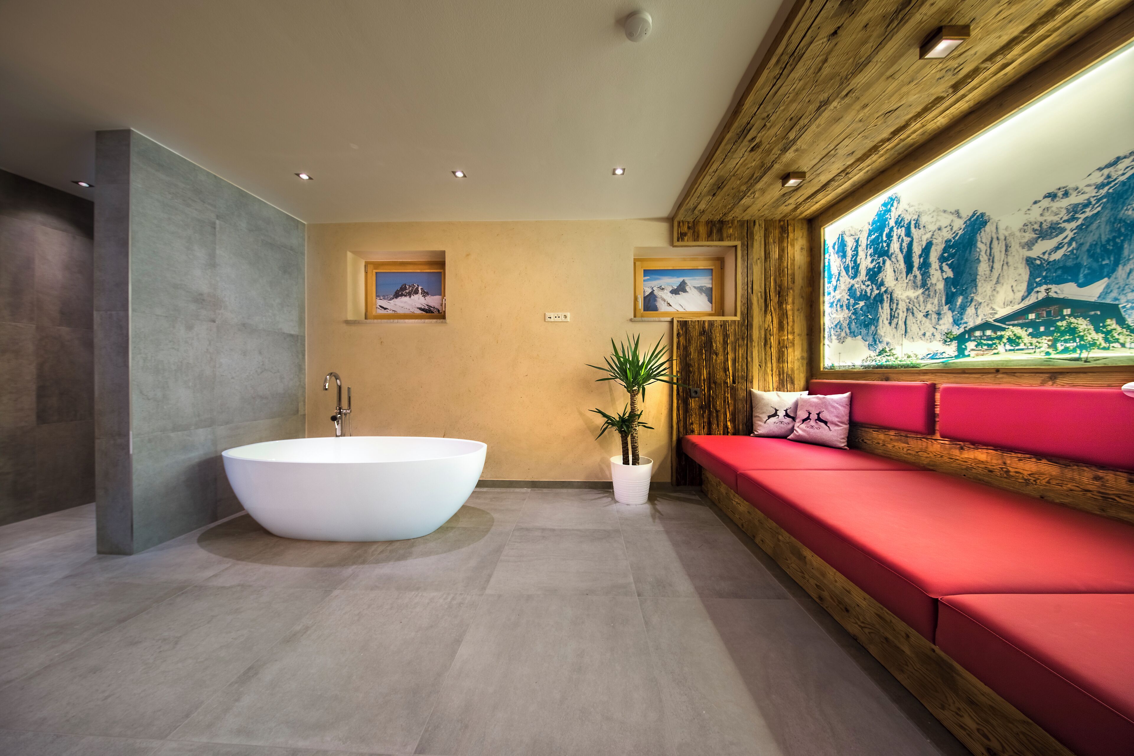 Un bagno moderno con una vasca freestanding, una panca di legno con cuscini rossi e un grande quadro appeso al muro raffigurante un paesaggio montano.