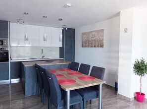A modern kitchen with white cabinets, a central island, and a dining area with a wooden table and blue chairs. - Dalmatia Apartments (Pakostane)