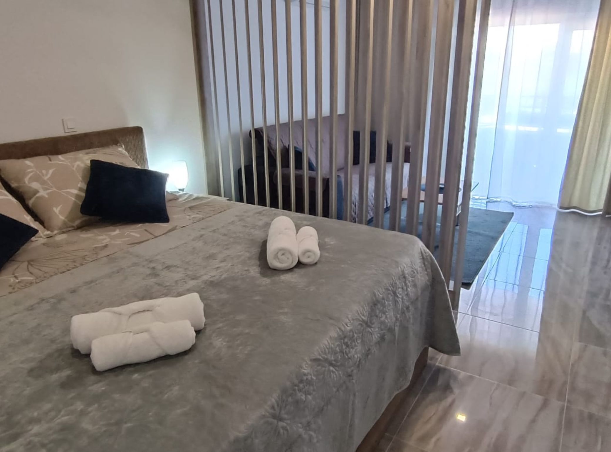 A neatly made bed with a grey bedspread and rolled towels, a wooden headboard, and a view of the outside through a window with vertical blinds.
