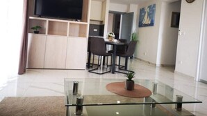 Un salon moderne avec une table basse en verre, un téléviseur à écran plat fixé sur un meuble en bois, un coin repas avec une table et des chaises, et une cuisine en arrière-plan.