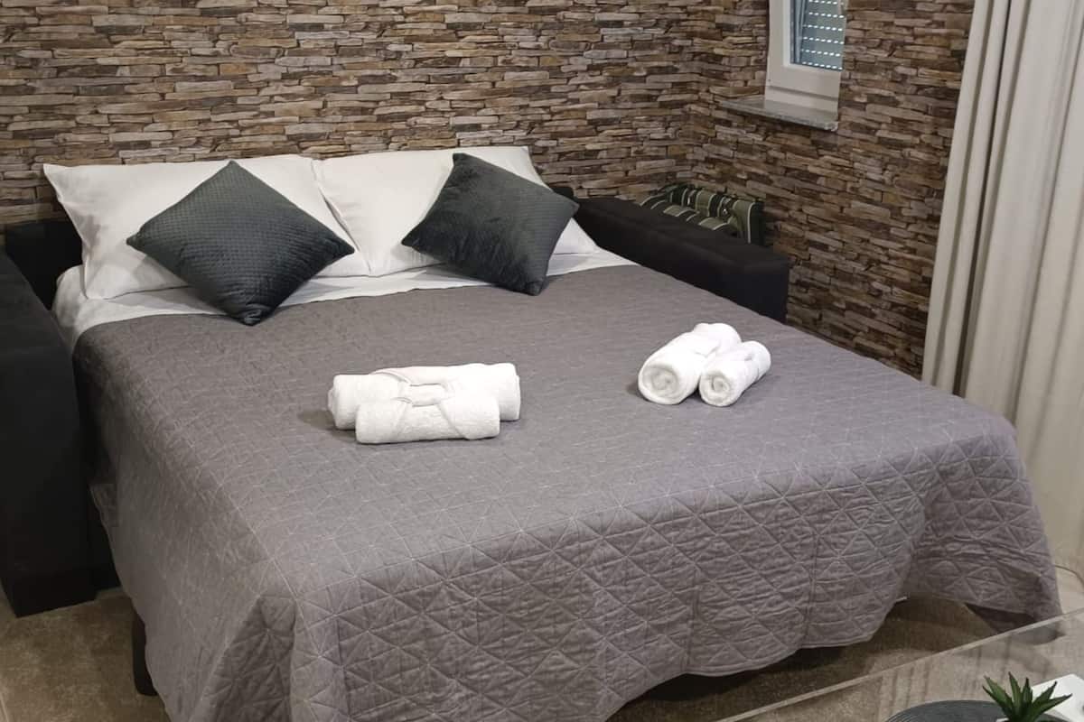 Un lit bien fait, recouvert d’une courtepointe grise matelassée, avec deux oreillers et deux serviettes enroulées posées sur le lit.