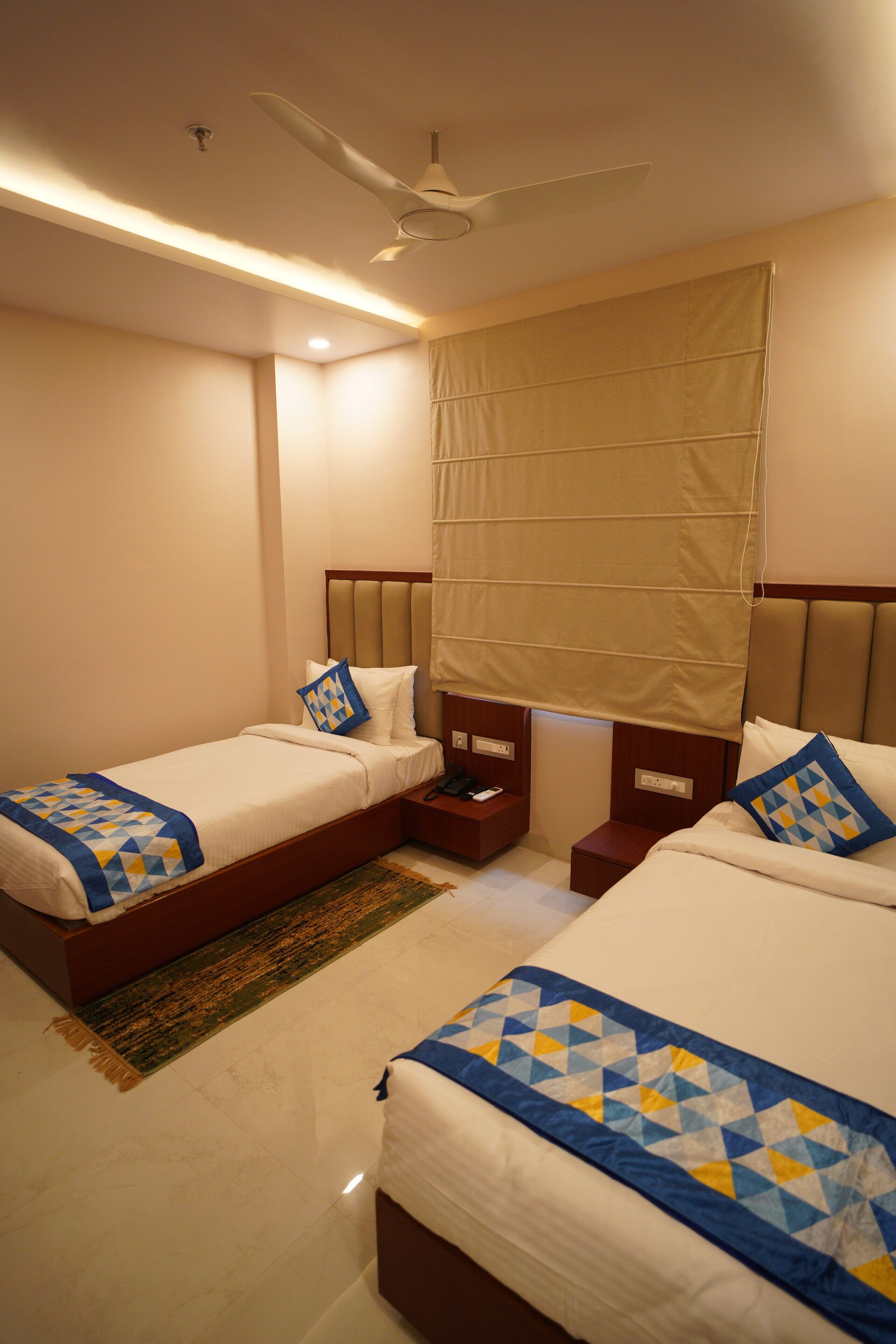 A hotel room with two beds, a ceiling fan, a window with curtains, and a small bedside table.