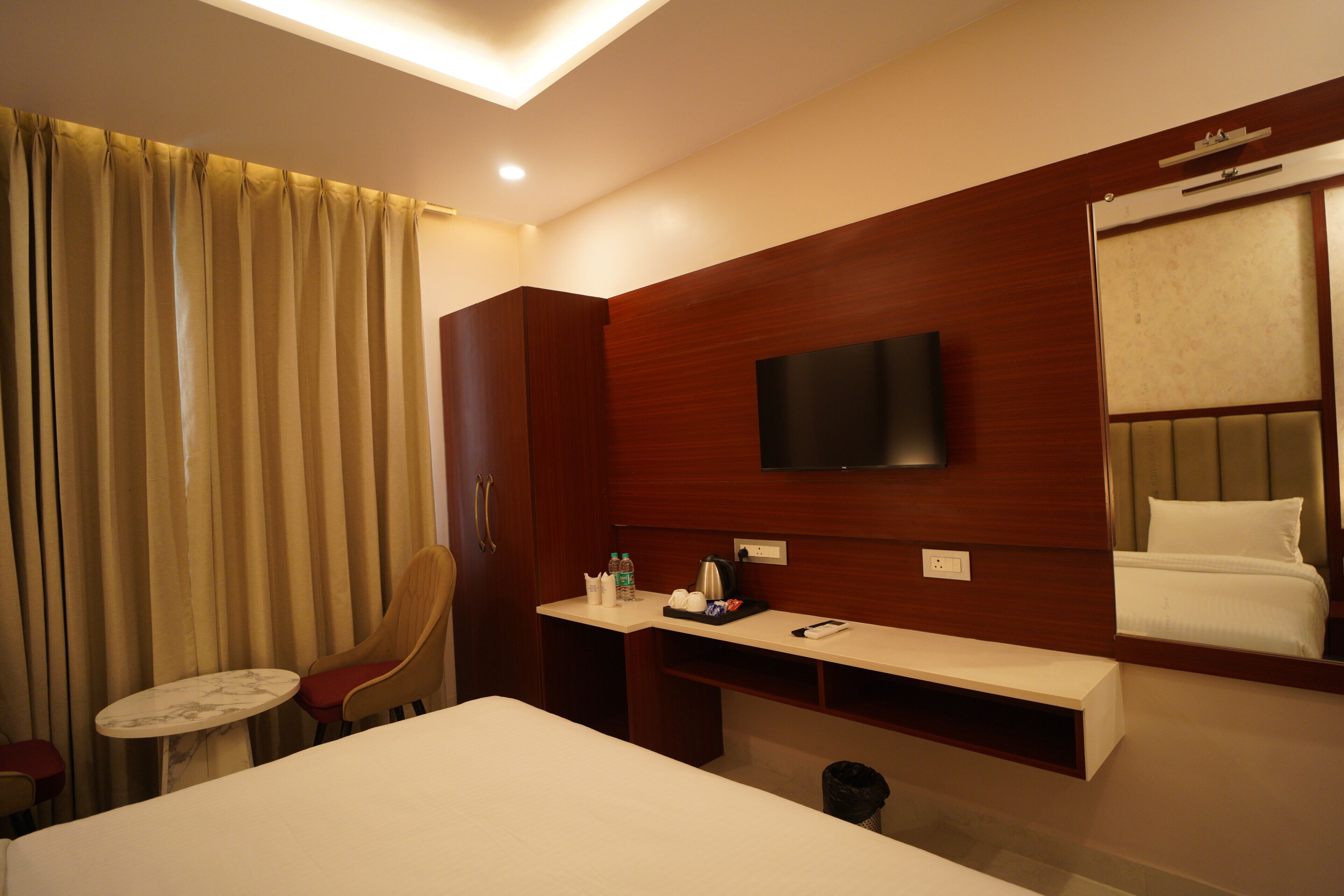A hotel room with a bed, a TV mounted on the wall, a desk with a chair, a small table, and a window with curtains.