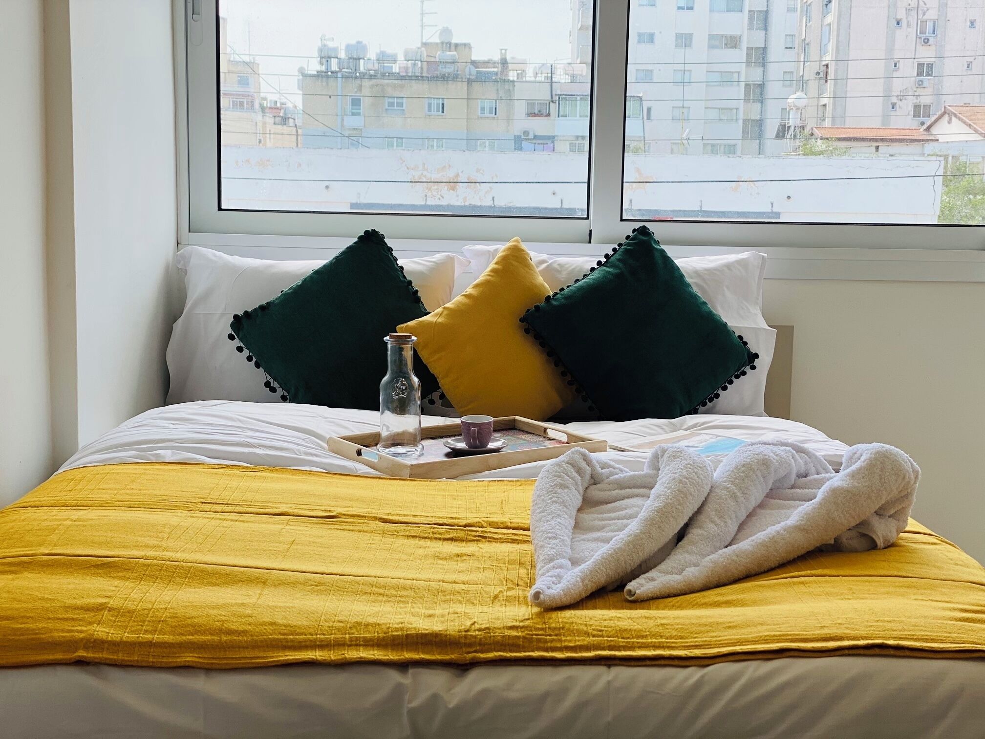 A neatly made bed with a yellow bedspread, two green and one yellow pillow, a glass bottle, a cup, and a folded towel on a tray.