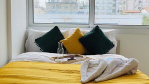 A neatly made bed with a yellow bedspread, two green and one yellow pillow, a glass bottle, a cup, and a folded towel on a tray. - Pink Flamingo 1-BR Apartment in Larnaca (Larnaca)