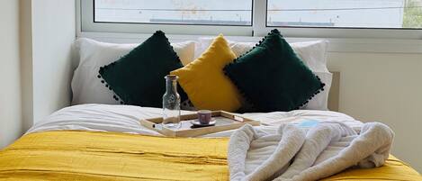 A neatly made bed with a yellow bedspread, two green and one yellow pillow, a glass bottle, a cup, and a folded towel on a tray.