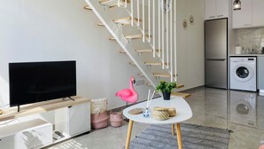 A modern living room with a flat-screen TV, a pink flamingo statue, a staircase with white railings, and a kitchen area with a washing machine. - Pink Flamingo 1-BR Apartment in Larnaca (Larnaca)