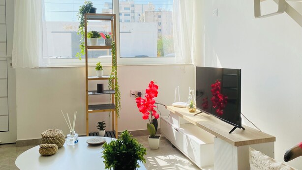 A modern living room with a large window, a TV on a wooden stand, a white sofa, a coffee table with decorative items, and a plant shelf. - Pink Flamingo 1-BR Apartment in Larnaca (Larnaca)