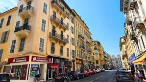 A street view with multi-story buildings, a row of parked cars, and storefronts including a real estate agency and a vape shop. - Big flat - Le Cala - City Center (Nice)