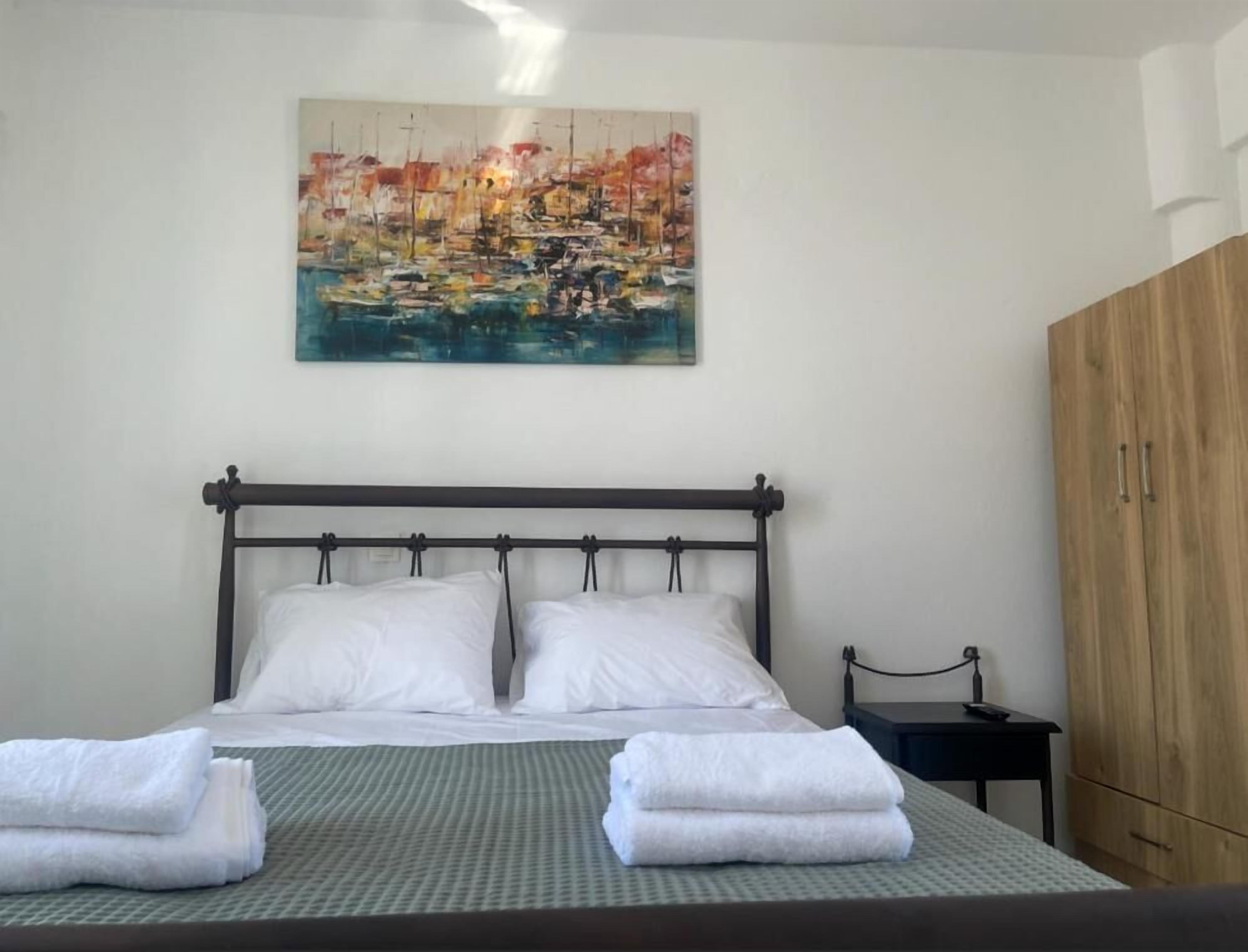 A neatly made bed with white linens and towels, a wooden headboard, and a painting of a harbor scene on the wall.