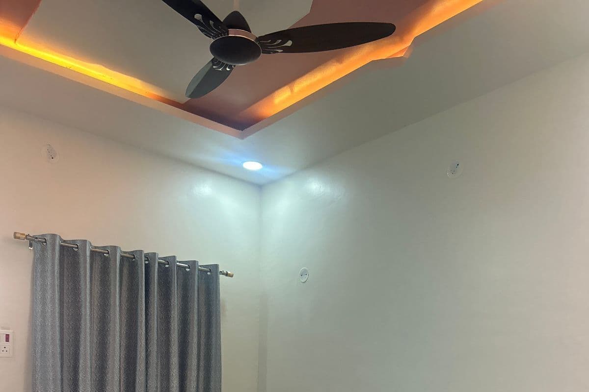 Une chambre à coucher avec un lit, deux oreillers, une tête de lit, un ventilateur de plafond et une fenêtre avec des rideaux.