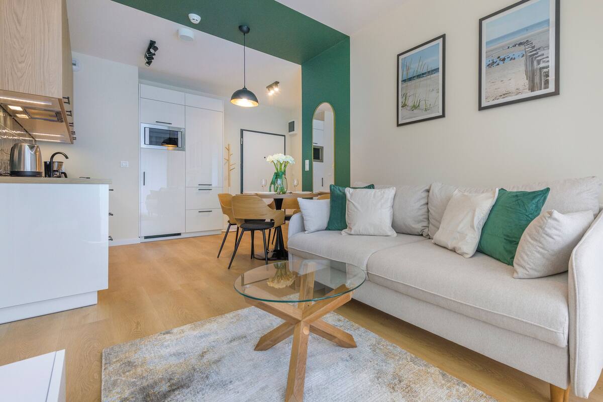Ein modernes Wohnzimmer mit Glastisch, einer Couch mit Kissen, einem Essbereich mit Stühlen und einer Küche im Hintergrund.