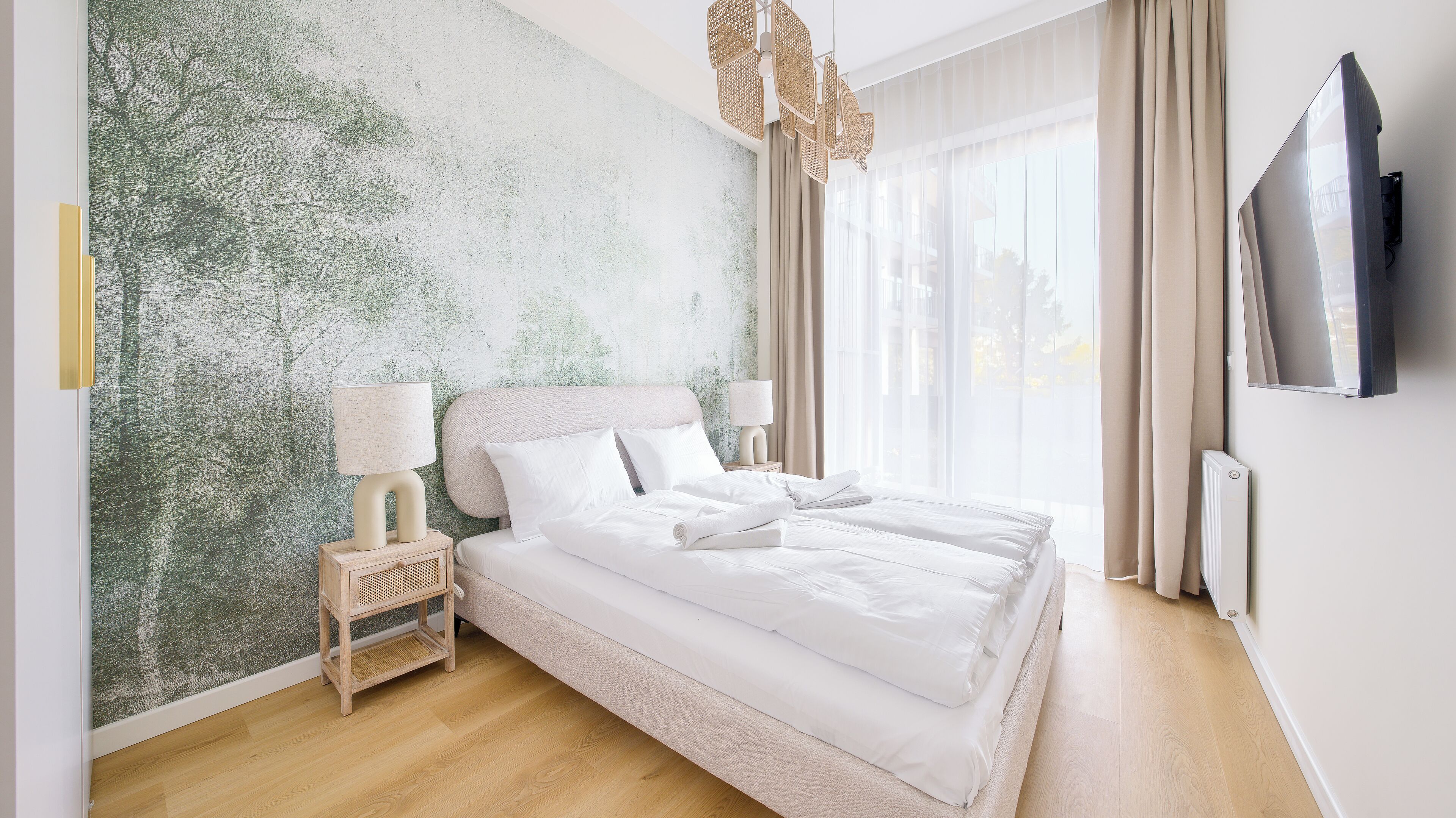 Una cama bien hecha con sábanas blancas, una mesita de noche con lámpara, un televisor montado en la pared y un gran ventanal con cortinas.