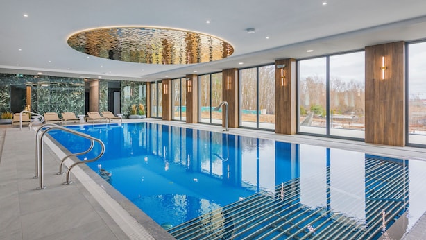 A modern indoor swimming pool with clear glass walls, lounge chairs, and a view of the outdoors. - Apartamenty Sun & Snow The One Ustronie Morskie (Sianożęty)