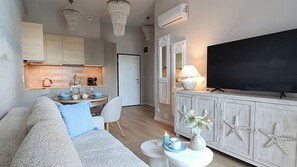 Un salon moderne comprenant un canapé, une table à manger, un téléviseur à écran plat et un coin cuisine avec des placards et un luminaire suspendu.
