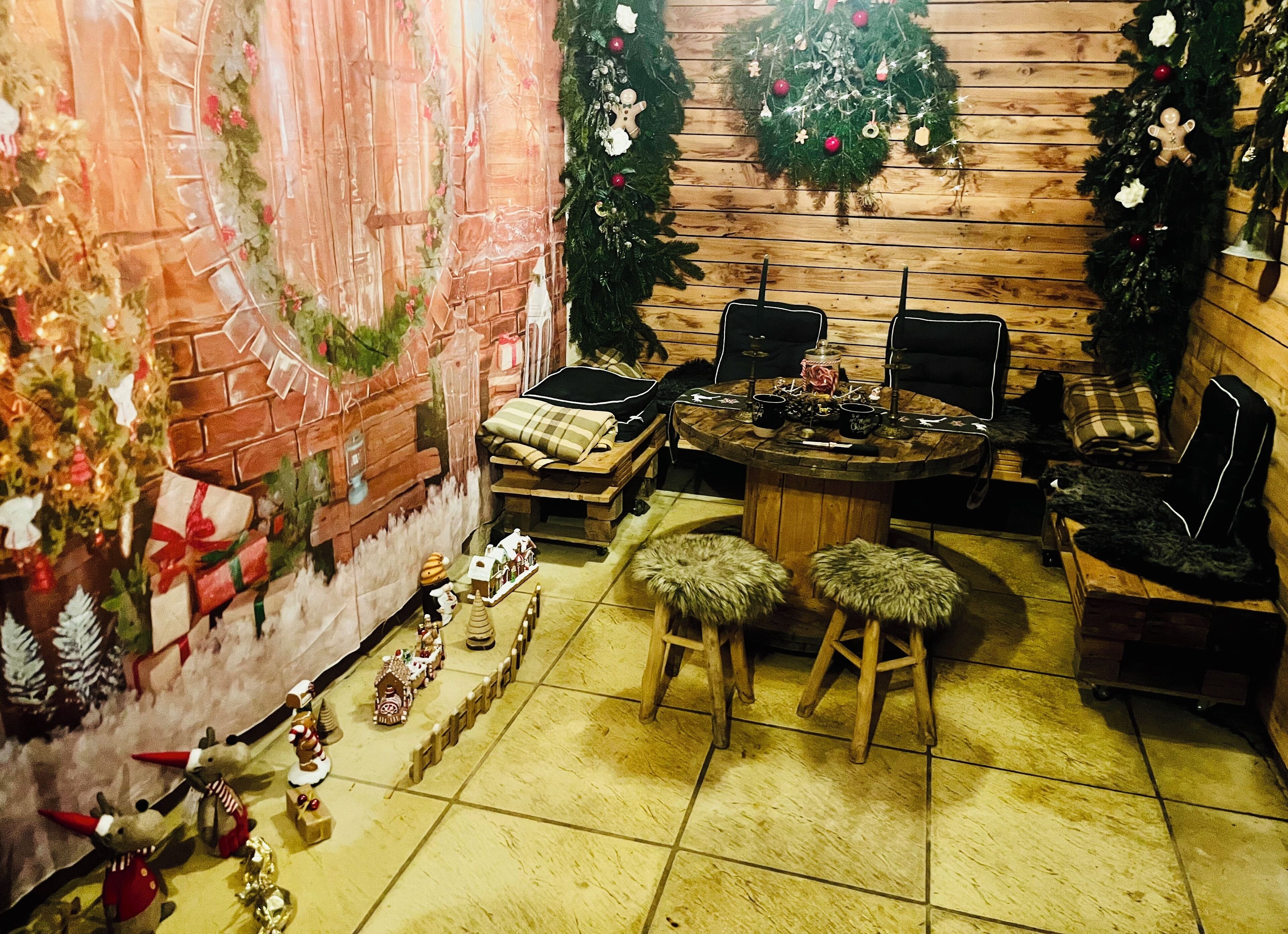 Ein gemütliches Zimmer mit Backsteinwand, Holzboden und einem runden Holztisch mit Stühlen. Weihnachtliche Dekorationen, darunter Kränze und ein Lebkuchenhaus, sind vorhanden.