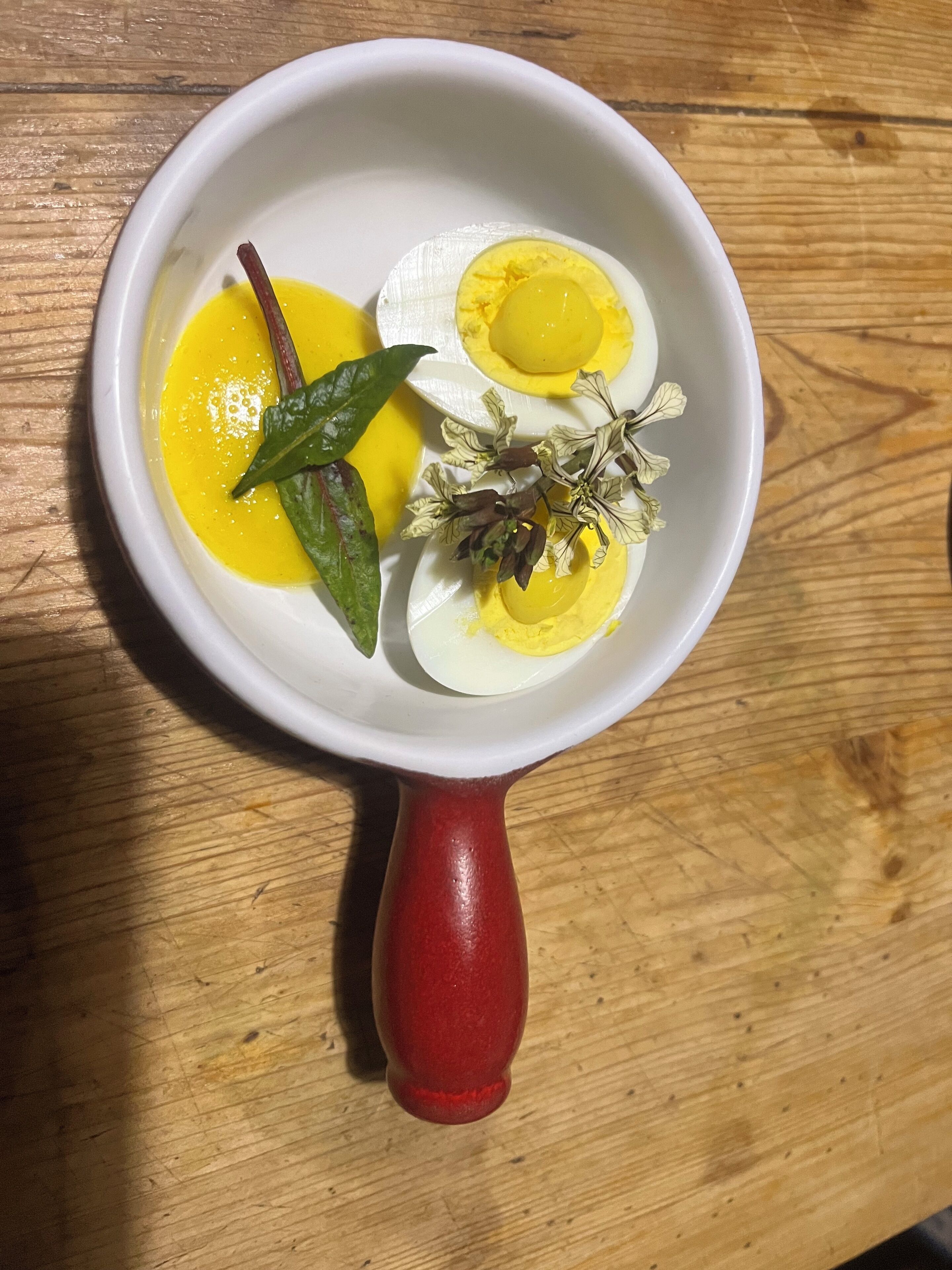 Een witte kom met een rode handgreep, gevuld met een gekookt ei, een chilipeper en gedroogde kruiden op een houten ondergrond.