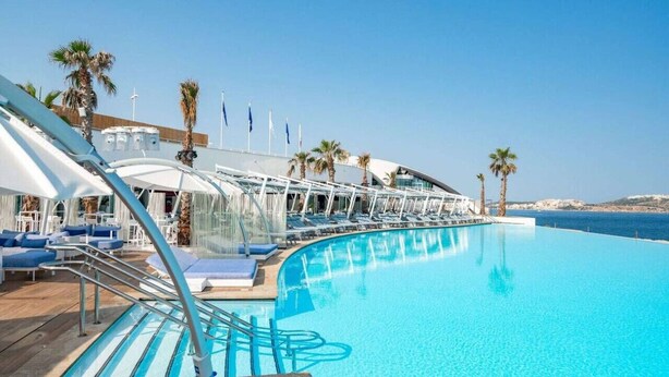 Area piscina di una nave da crociera, con poltronne prendisole e ombrelloni, una piscina azzurra e una vista sul mare.