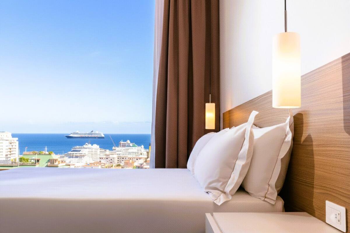 Ein Hotelzimmer mit Bett, Nachttisch, Lampe und Blick auf das Meer und Schiffe.