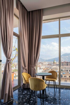 Una habitación con un ventanal grande, dos sillas amarillas, una mesa redonda y vistas a un paisaje urbano con montañas.