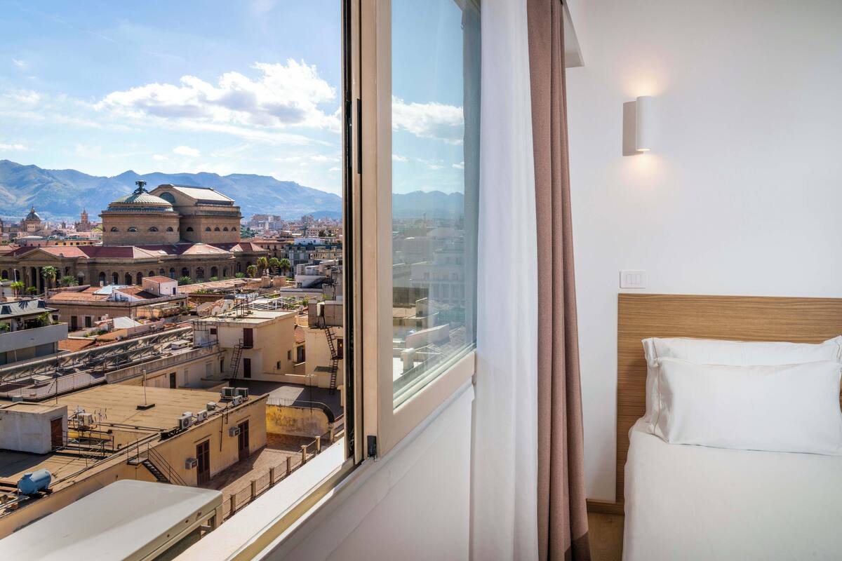 Ein Hotelzimmer mit großem Fenster und Blick auf ein historisches Gebäude und Berge.
