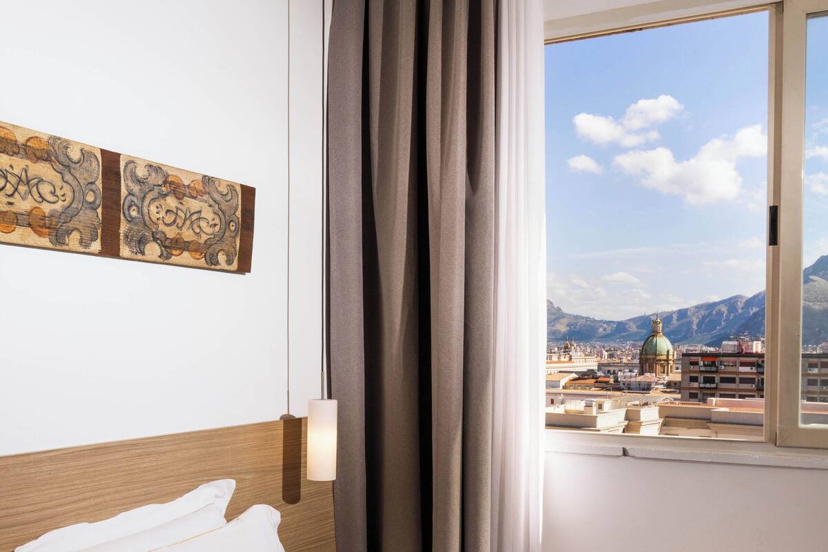 Ein Hotelzimmer mit Bett, Nachttisch, Lampe und Fenster mit Blick auf eine Stadtlandschaft und Berge.