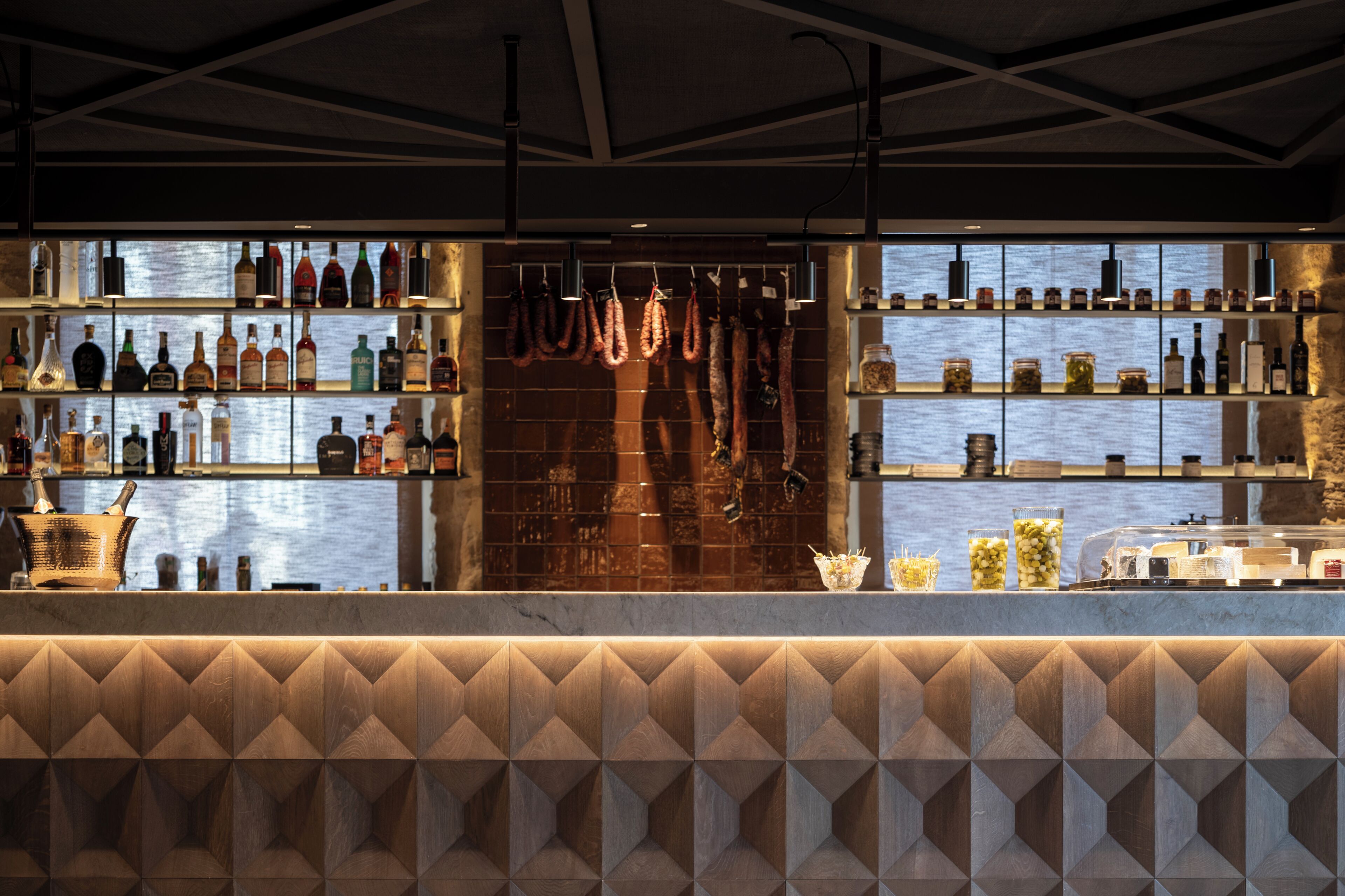 A bar area with shelves of bottles and jars, a patterned wall, and a view of the water.