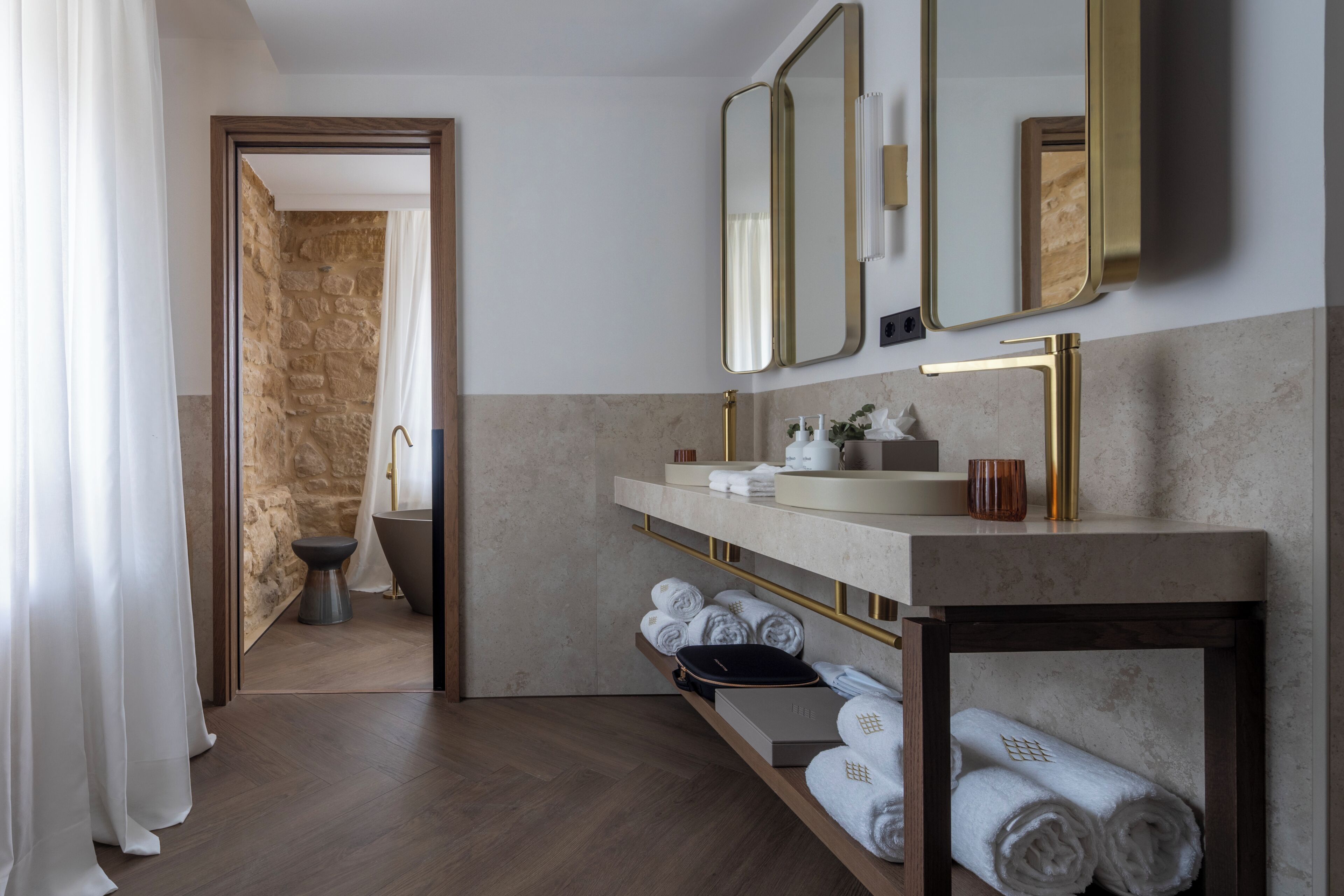 A modern bathroom with a stone wall, a bathtub, a vanity with a gold faucet, and a mirror.