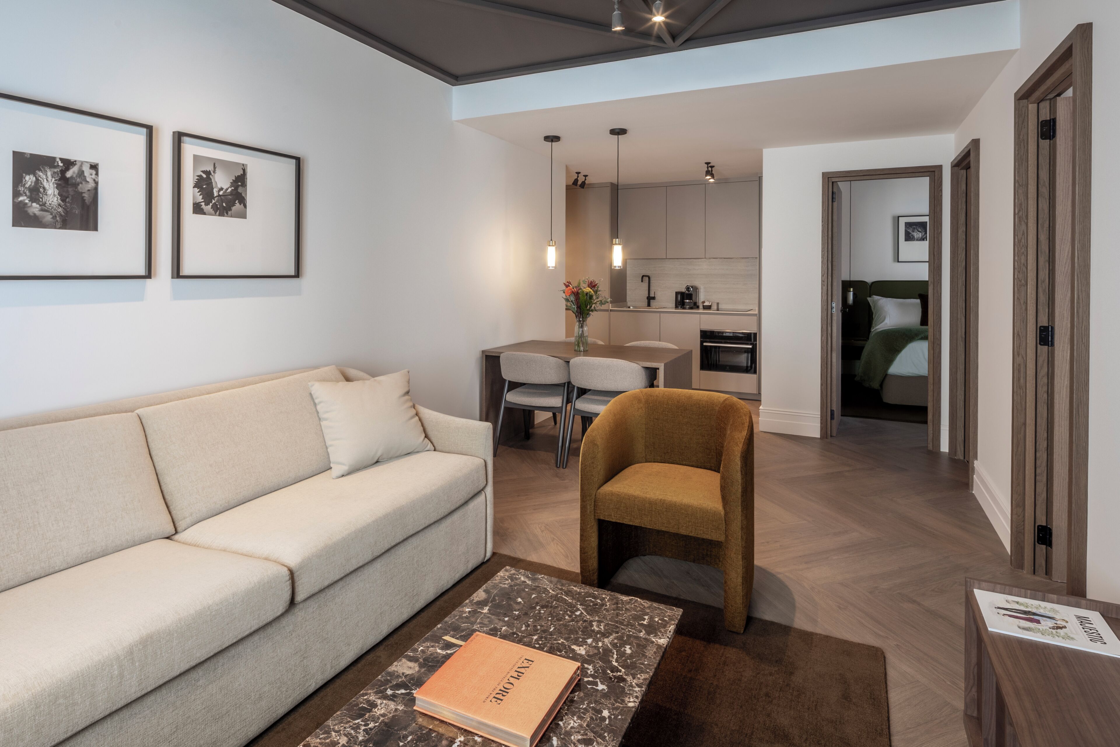 Una sala de estar moderna con un sofá, una mesa de centro con un libro, una zona de comedor con mesa y sillas, y una cocina al fondo.