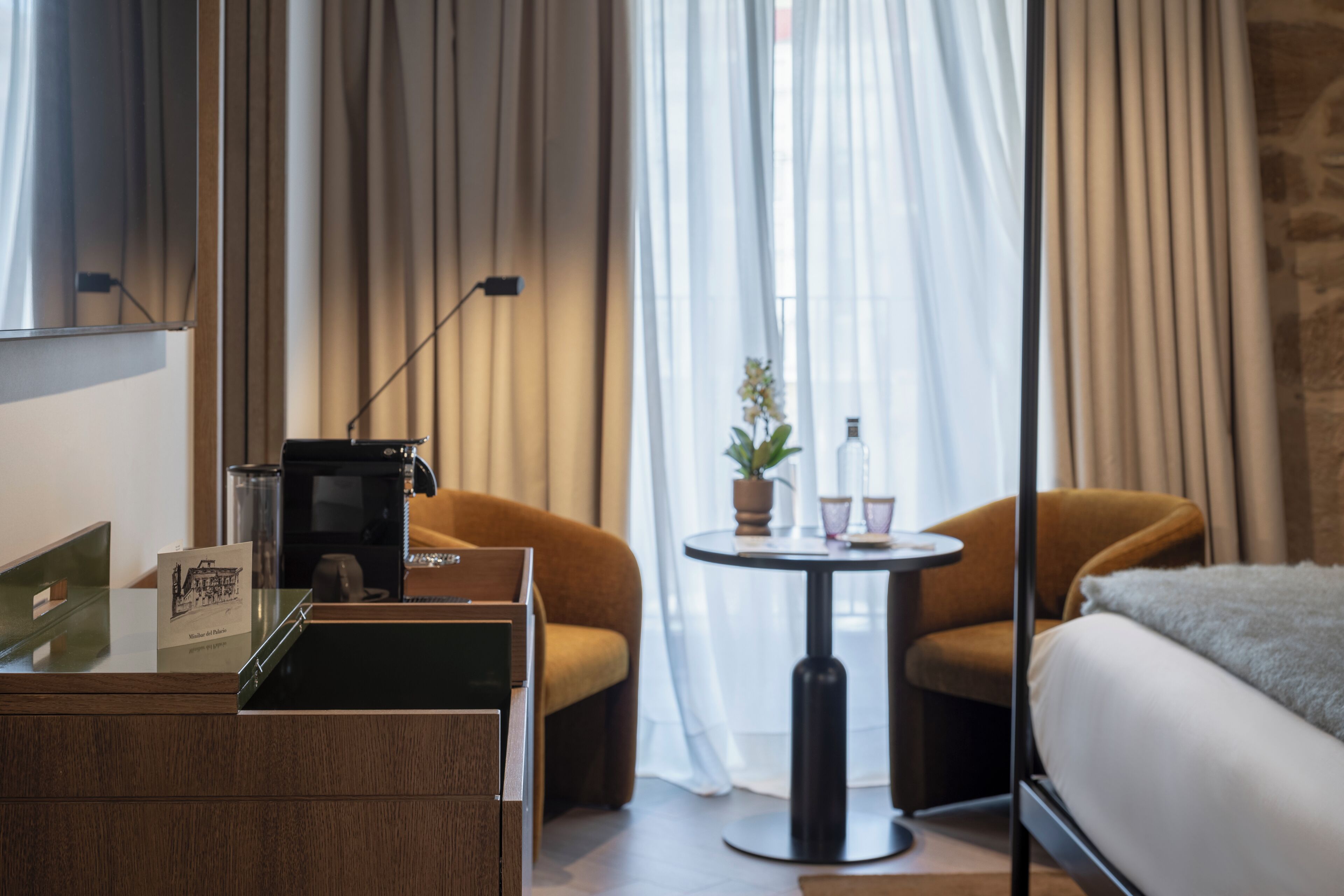 A hotel room with a bed, two armchairs, a small round table, a coffee machine, and a potted plant.