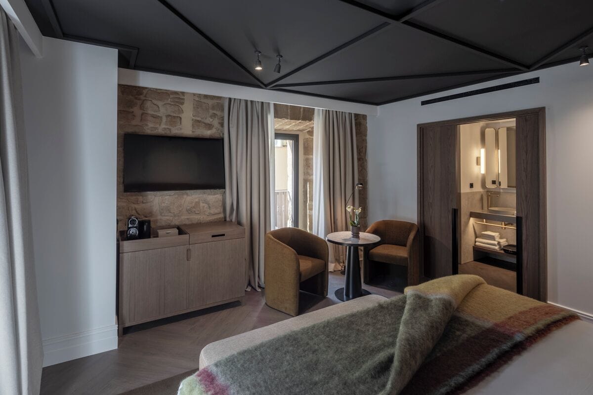 Una habitación de hotel moderna con una pared de acento de piedra, un televisor de pantalla plana, una pequeña mesa redonda, dos sillones, un sofá y un vestidor.