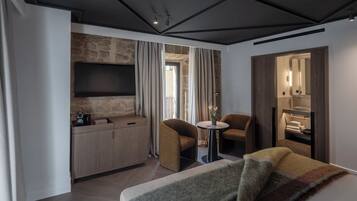 A modern hotel room with a stone accent wall, a flat-screen TV, a small round table, two armchairs, a sofa, and a walk-in closet.